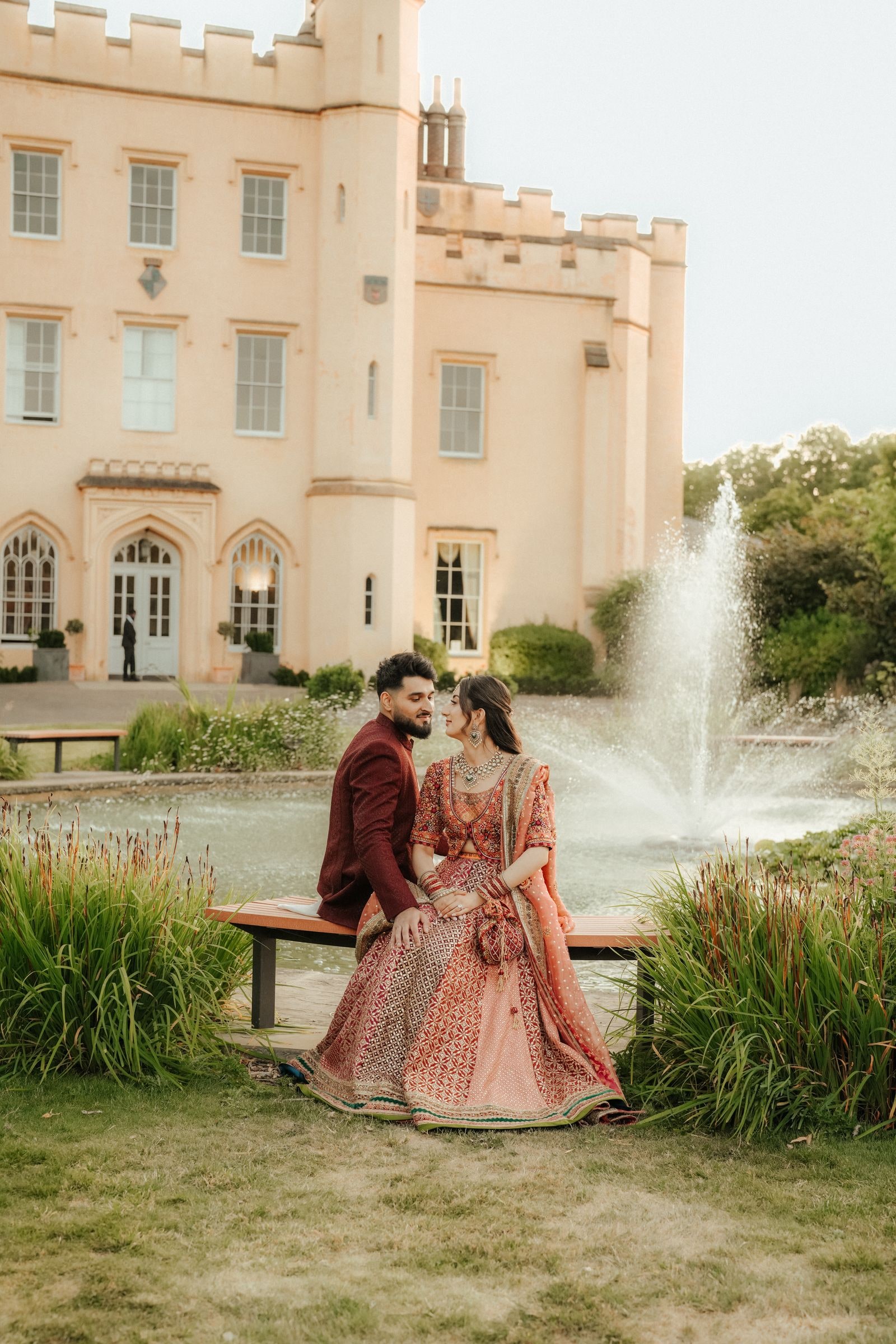 Couple in red and burgundy mehndi outfits on a bench beside the fountain at Ditton Manor