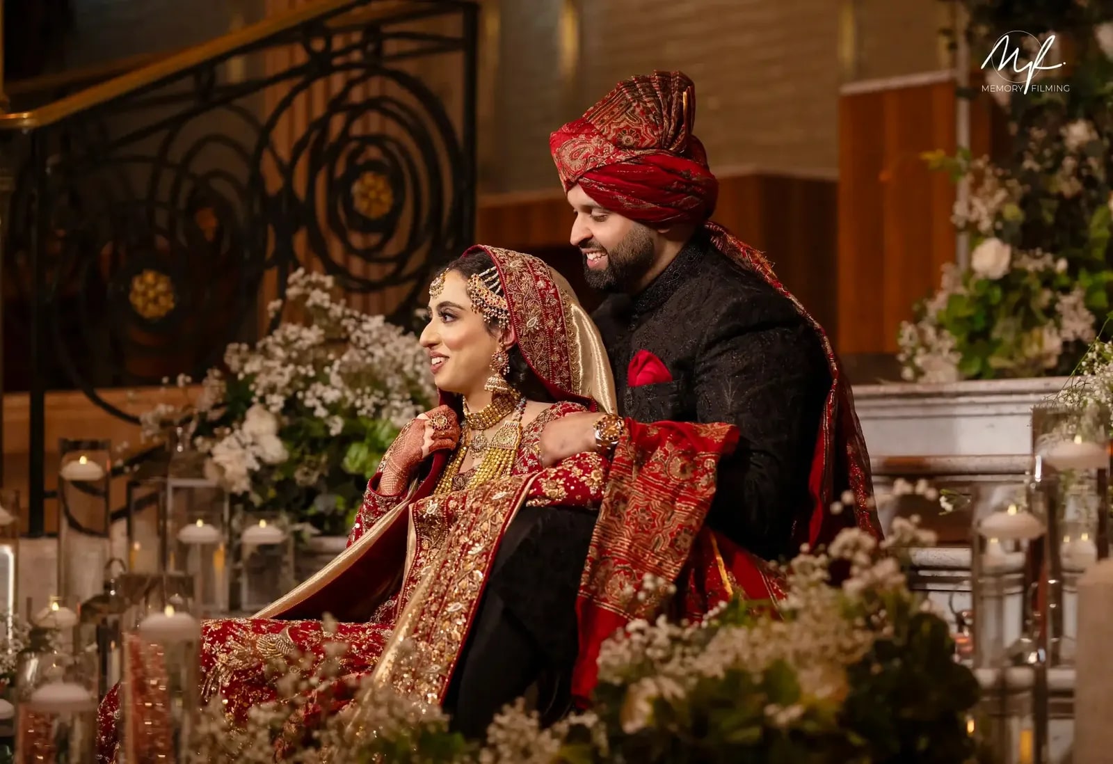 Bride and groom seated among candles and white flowers, he in a red turban and black sherwani, she in a red and gold lehenga
