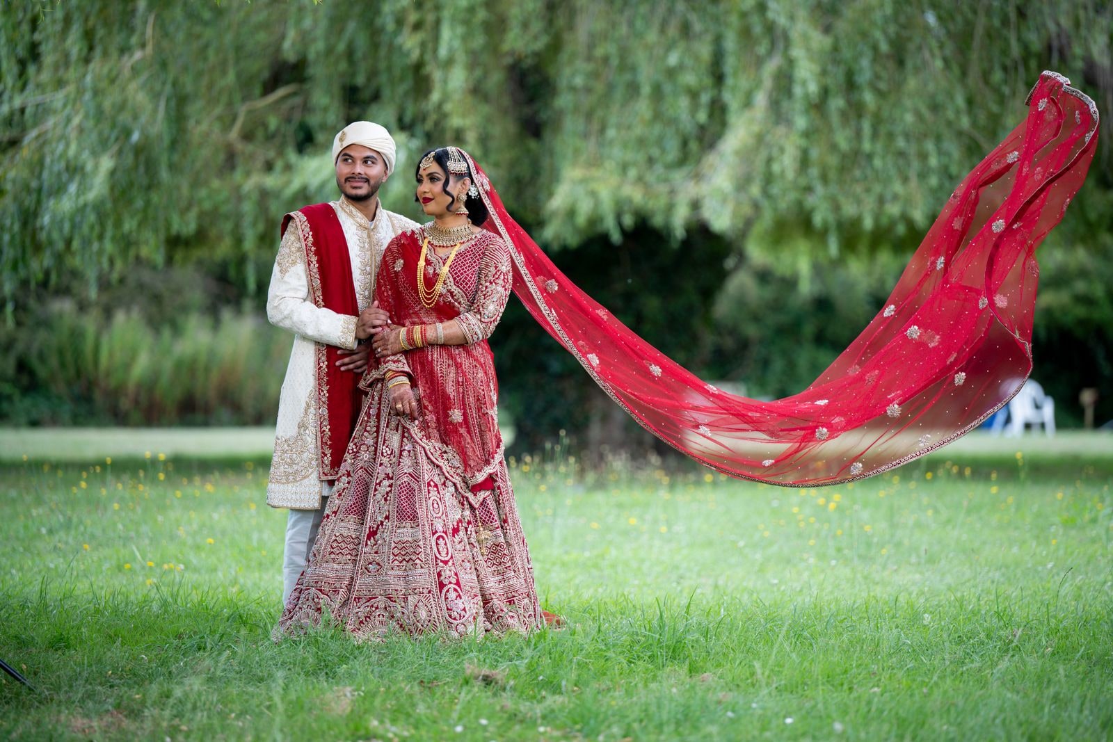 Bengali bride in a red lehenga with her dupatta catching the wind, beside her groom in an ivory sherwani