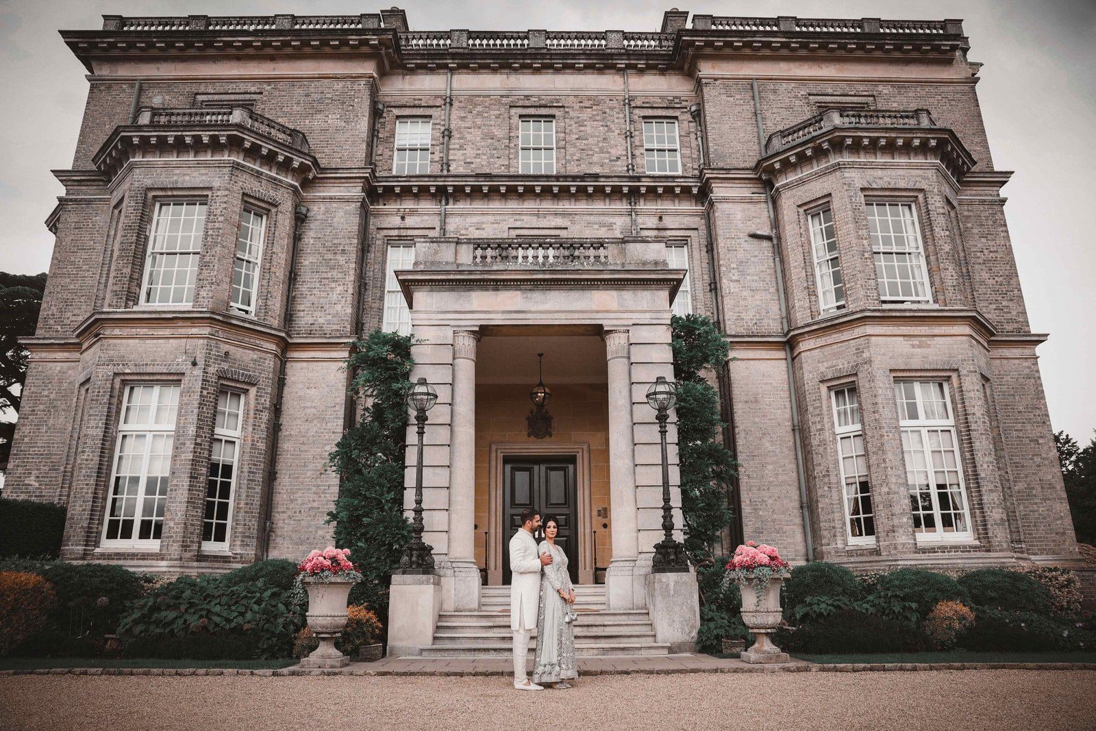 Couple standing small in an asian wedding venues london Hedsor House, the grey stone front rising above them.