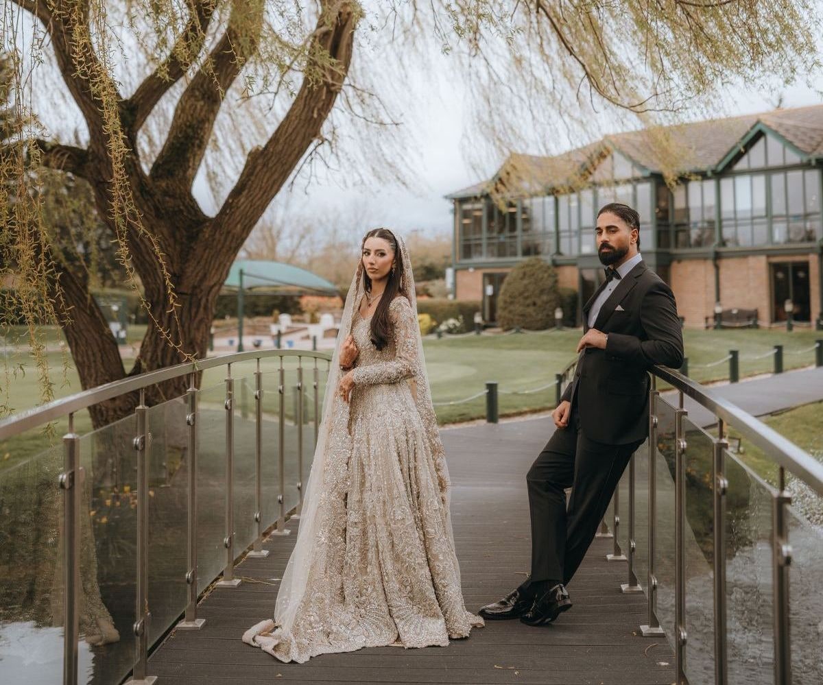 Muslim bride in a pale embroidered gown and groom in a dark suit on a footbridge over the lake at Three Rivers for their walima shoot.