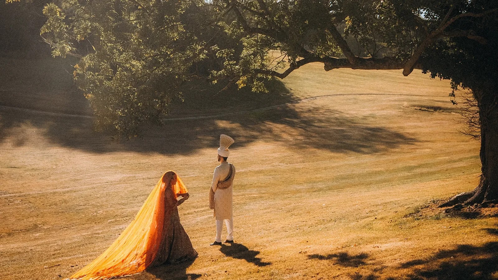 Muslim bride for nikkah phototgraphy in a gold and orange dupatta with a long train facing her groom in a cream sherwani and turban, in a sunlit field